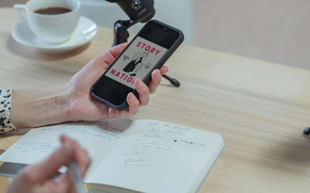 Žena sedi za radnim stolom na kojem se nalazi sveska i gleda u svoj telefon