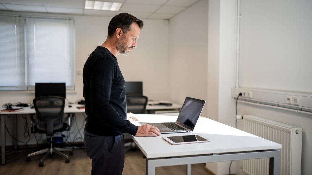 Muškarac stoji za stolom i radi na laptopu u kancelariji